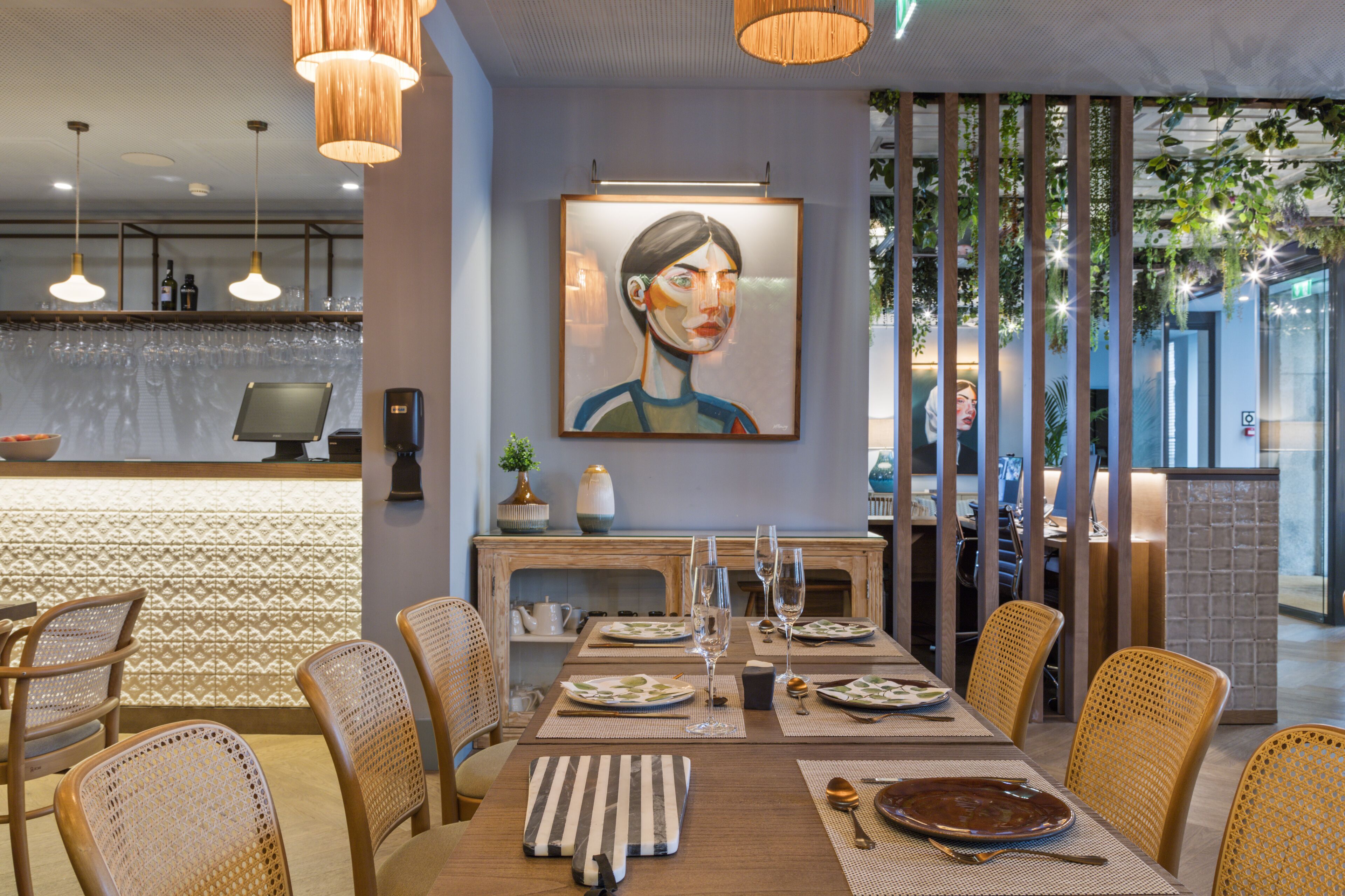 A modern dining area with a long table, wicker chairs, and a painting on the wall.