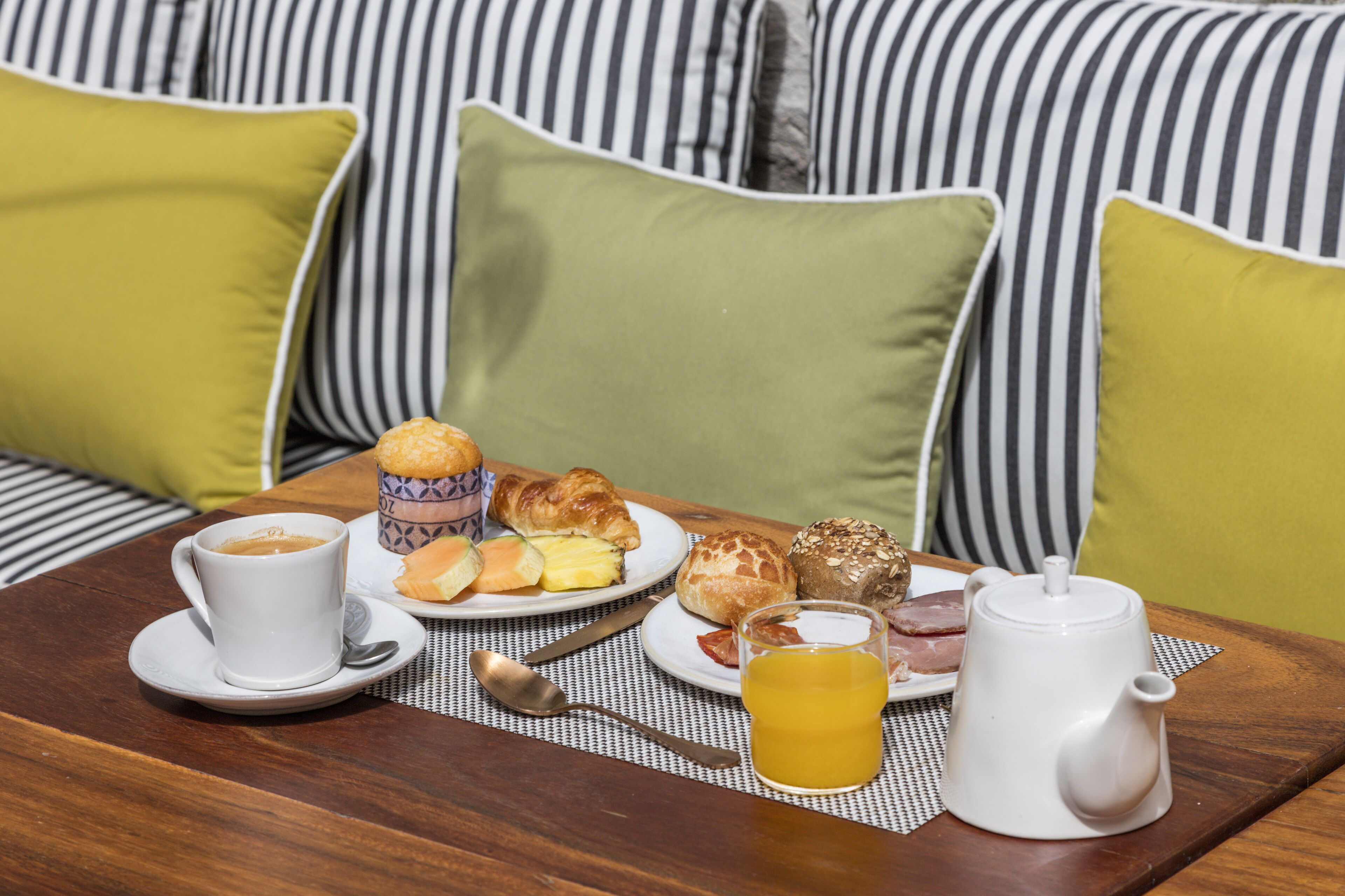 A breakfast table with coffee, croissants, muffins, and juice, set on a wooden table with a striped cushion in the background.