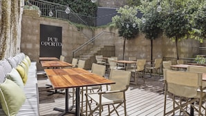 Un patio al aire libre con mesas y sillas de madera, un muro de piedra y una escalera que sube.