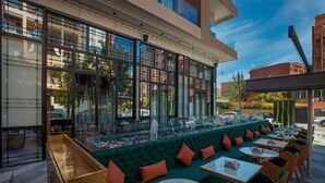 An outdoor seating area with green cushioned benches and round tables, a glass enclosure with a view of the street, and a modern building facade. - Stars Hotel (Marrakech)