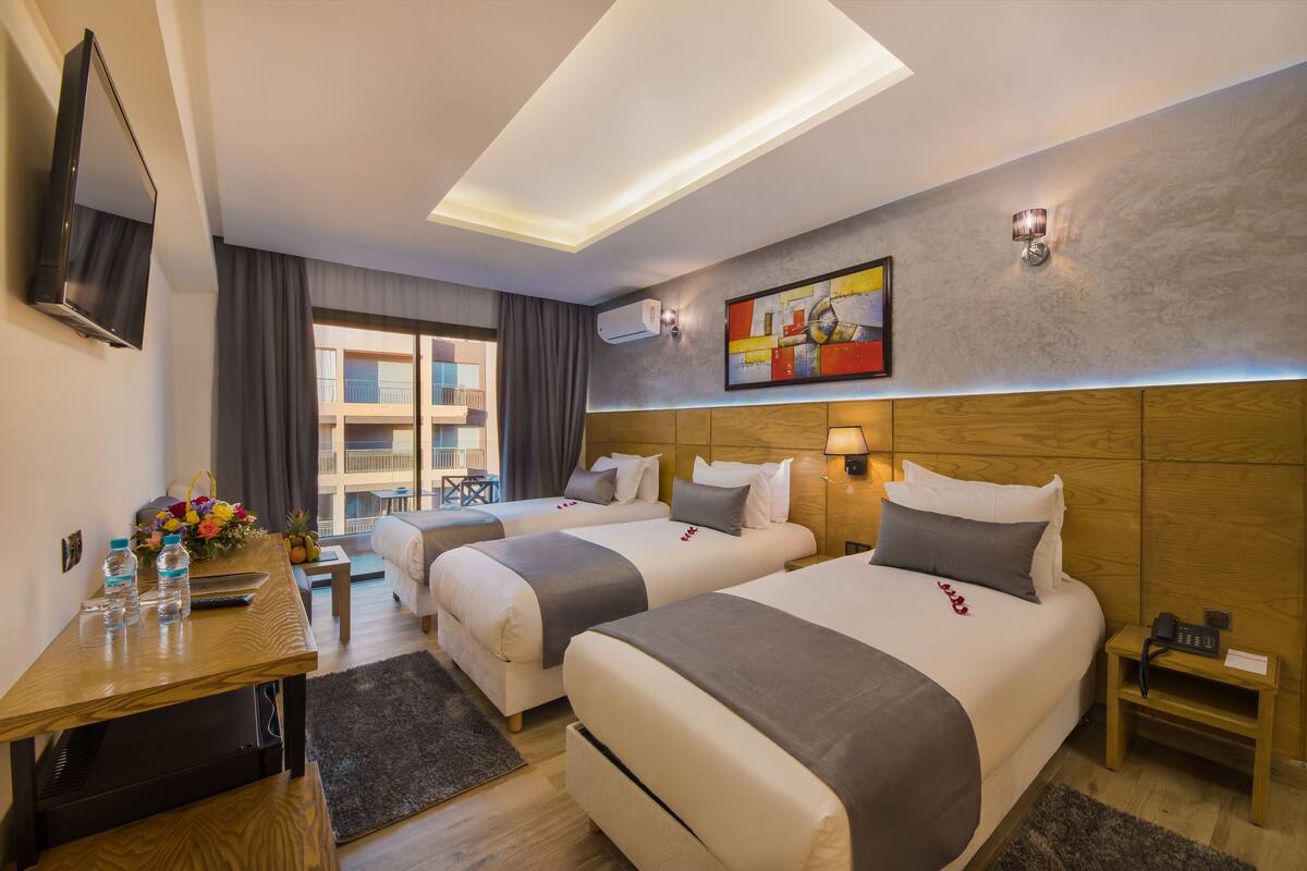 Ein Hotelzimmer mit zwei Betten, einem Schreibtisch mit Flachbildfernseher, einem kleinen Tisch mit Blumenvase und einem Bild an der Wand.