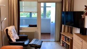 A living room with a black leather sofa, a small ottoman, a bookshelf, a television, and a sliding glass door leading to an outdoor area. - Apartment with loggia in a quiet complex, about 10 minutes walk to the (Holm)