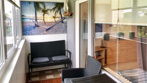 A balcony with a wicker sofa, a small table, and a glass door leading to a room with a wooden cabinet and a television. - Apartment with loggia in a quiet complex, about 10 minutes walk to the (Holm)