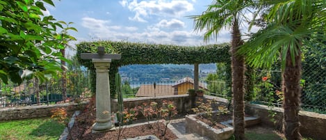 A garden with a stone pillar, a pergola covered in greenery, and a view of a distant town.