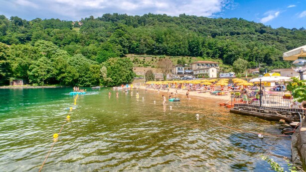 A lakeside beach with clear water, surrounded by lush green hills.