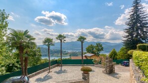 A paved courtyard with palm trees, a stone planter, and a view of mountains and a lake.
