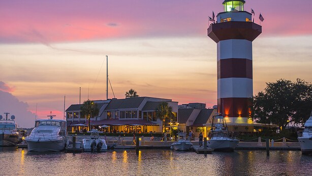 Un faro al atardecer con barcos amarrados cerca y un edificio con mesas al aire libre.