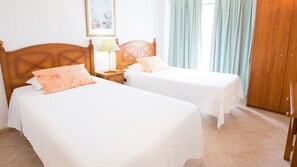 A hotel room with two single beds, a wooden headboard, a nightstand with a lamp, a painting on the wall, and a wooden wardrobe. - Apartamentos Quinta da Praia (Portimão)