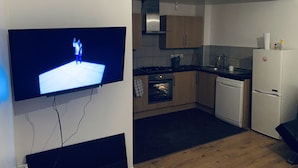 A kitchen with a flat-screen TV mounted on the wall, wooden cabinets, a stove, and a refrigerator. - 2-bed Apartment (Kingston Upon Thames)
