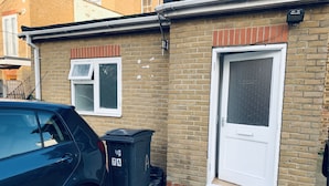 A black car parked in front of a house with a white door and a window with a white frame. There is a black trash bin next to the car. - 2-bed Apartment (Kingston Upon Thames)
