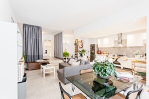 A modern living room with a glass dining table, grey sofa, and a kitchen area with white cabinets and appliances.