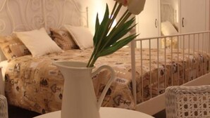 Une pièce avec un lit, une table blanche sur laquelle se trouve une carafe, et une chaise en osier.