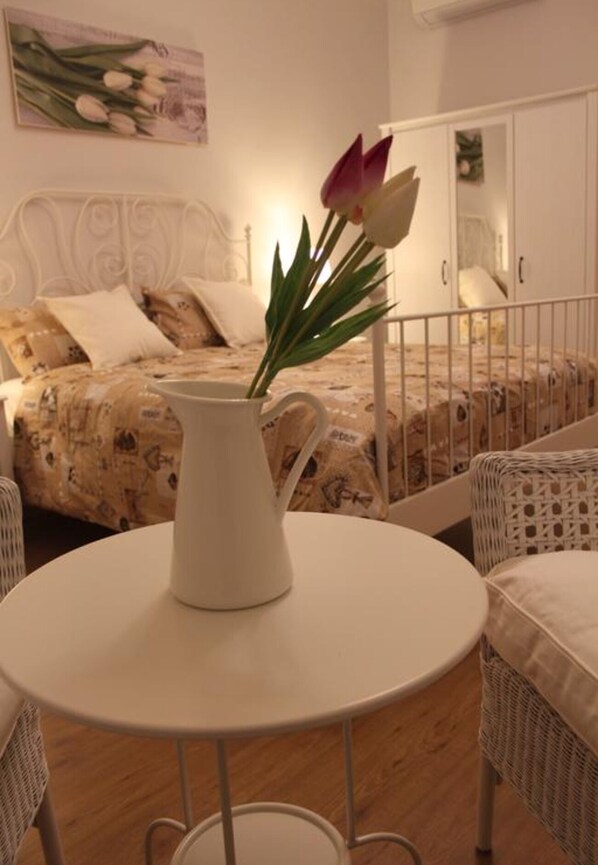 A room with a bed, a white table with a pitcher, and a wicker chair.