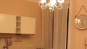 Une cuisine compacte avec des meubles blancs, une cuisinière à gaz, un évier et une table à manger avec des chaises. Un lustre est suspendu au-dessus de la table, et une horloge est visible au mur.