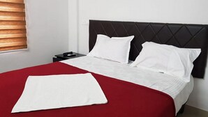 A neatly made bed with a red bedspread and white pillows, a black headboard, and a bedside table with a remote control. - Thulasi Inn (Kanayannur)