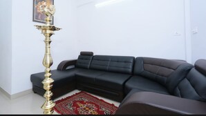 A living room with a black sectional sofa, a decorative brass floor lamp, a red patterned rug, and a framed picture on the wall. - Thulasi Inn (Kanayannur)