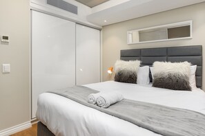 A modern bedroom with a large bed, grey headboard, fluffy pillows, and a bedside table with a lamp.