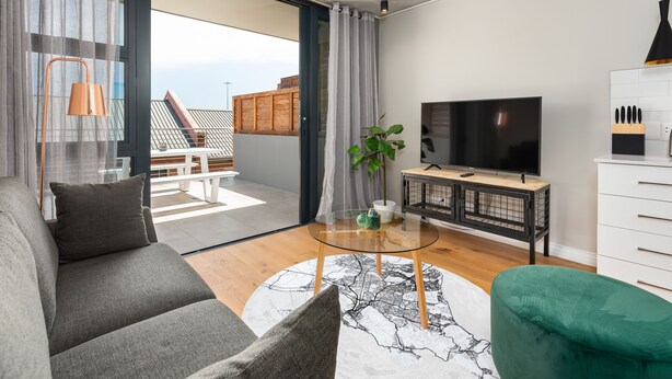 Ein modernes Wohnzimmer mit einer Couch, einem Couchtisch und einem Fernseher. Ein Balkon bietet Blick auf den Himmel und die Stadt.