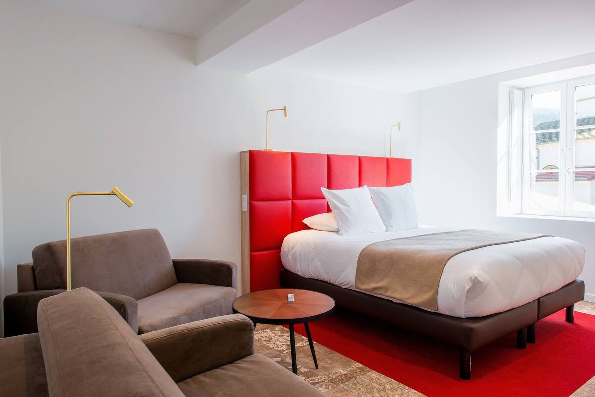 A modern hotel room with a red upholstered headboard, a bed with white linens, a brown sofa, a small wooden side table, and a large window providing a view.