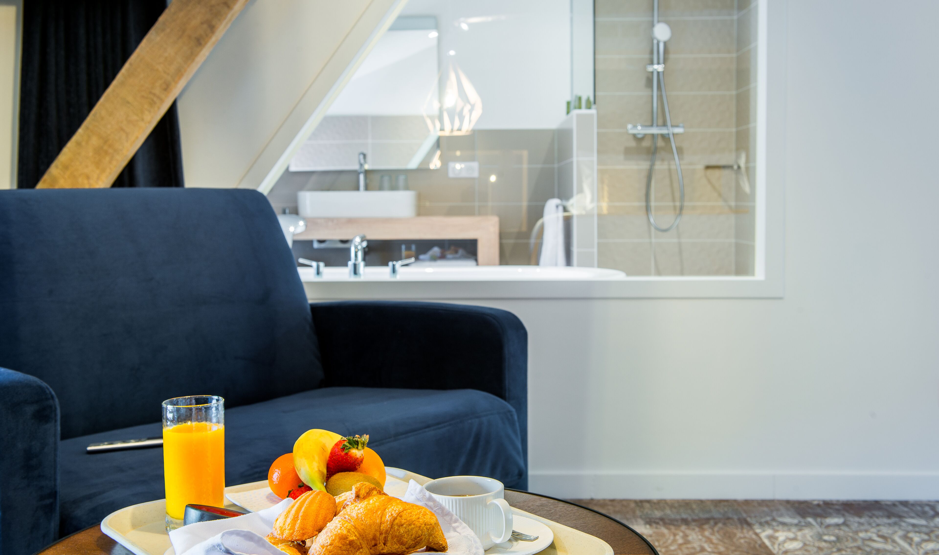 Un plateau de petit-déjeuner avec des croissants, des fruits, du jus d’orange et du café est posé sur une table dans une chambre moderne, où l’on aperçoit un canapé bleu et, par la fenêtre, une salle de bain avec une douche.