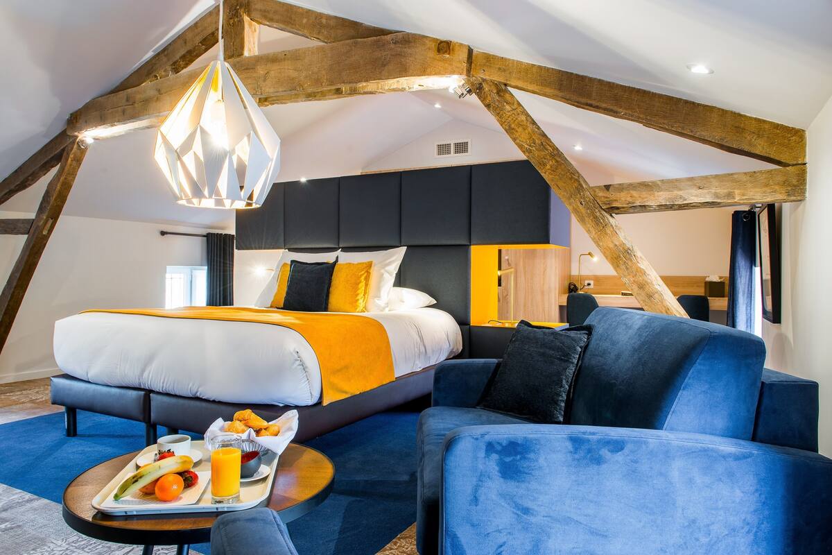 A modern hotel room with a large bed, a blue sofa, and a breakfast tray with fruits and juice.