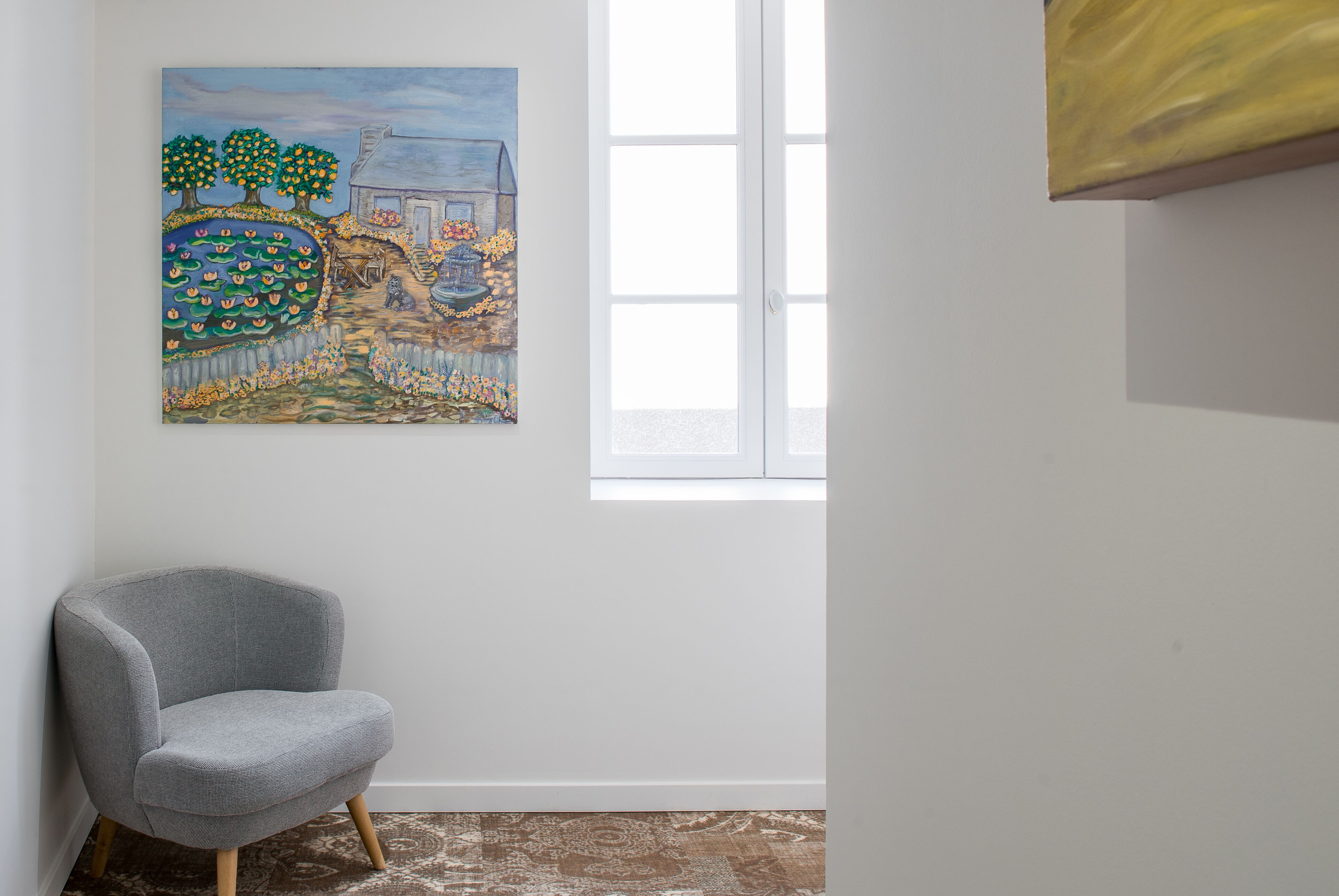 Une pièce avec un fauteuil gris, un tableau représentant un paysage et une fenêtre avec des stores.