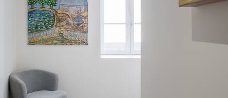 Une pièce avec un fauteuil gris, un tableau représentant un paysage et une fenêtre avec des stores.