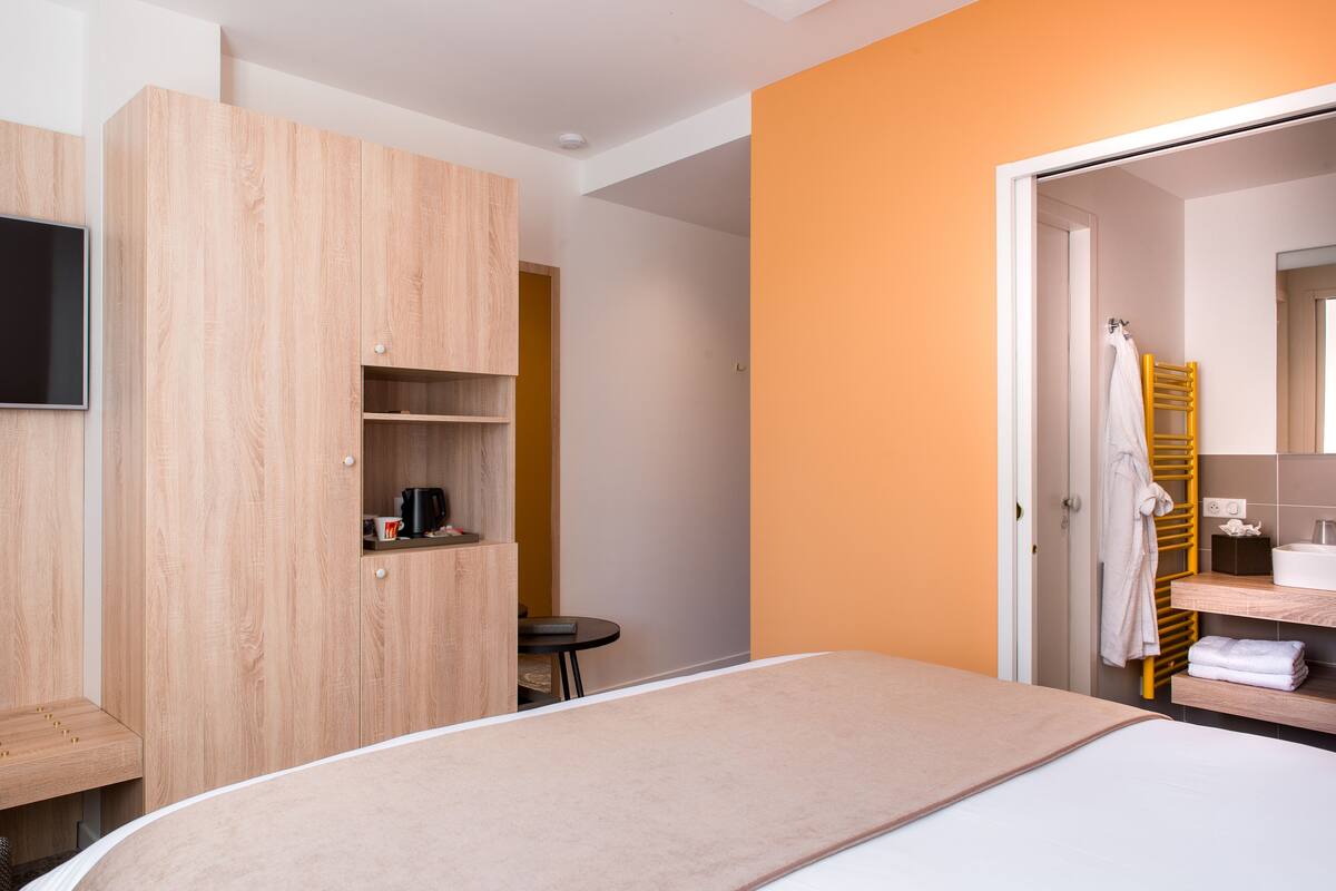 A hotel room with a bed, a wooden wardrobe, a small table with a coffee maker, and a bathroom with a yellow towel rack.