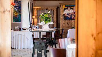 Un área de comedor con mesas dispuestas para dos, un jarrón blanco con flores y un cuadro en la pared.