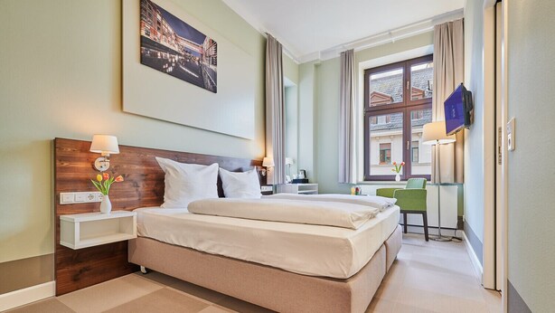 A hotel room with a large bed, a bedside table with a lamp, a small desk with a chair, and a window with curtains. - Inklusionshotel PHILIPPUS Leipzig (Leipzig)
