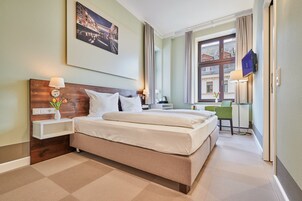 Una habitación de hotel con una cama grande, una mesita de noche con lámpara, un escritorio pequeño con silla y una ventana con cortinas.