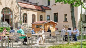 An outdoor café with tables and chairs arranged in rows, set against a building with arched windows and a small wooden structure in the background. - Inklusionshotel PHILIPPUS Leipzig (Leipzig)