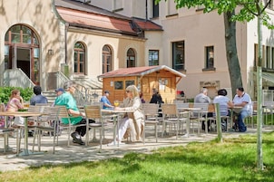 Un café al aire libre con mesas y sillas dispuestas en filas, frente a un edificio con ventanas de arco y una pequeña estructura de madera al fondo.