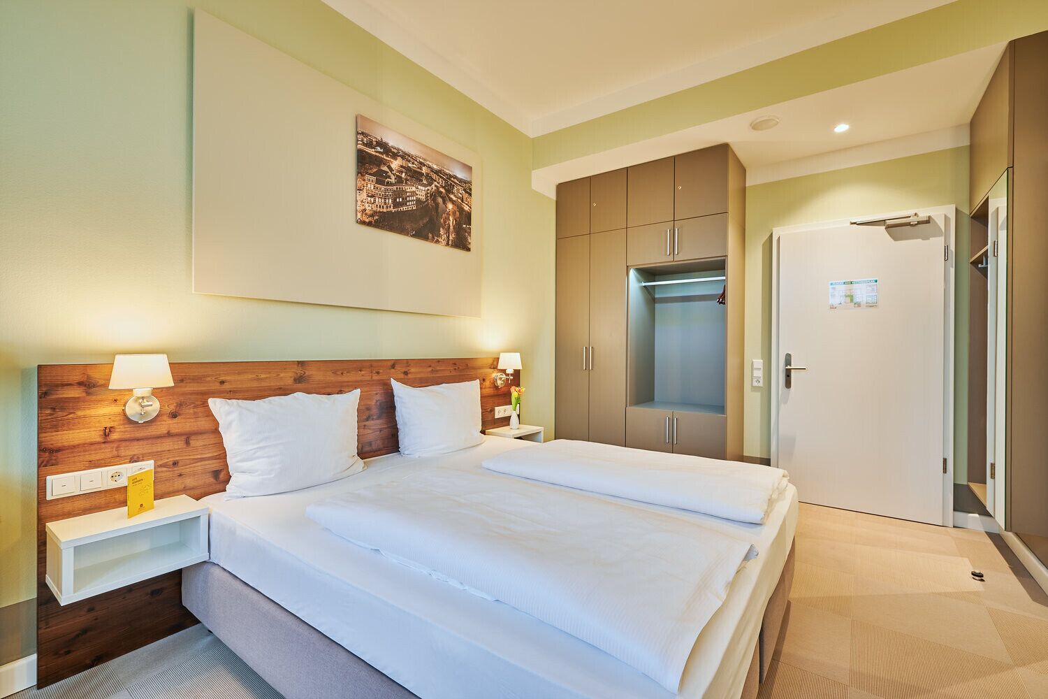 Ein Hotelzimmer mit Doppelbett, einem Holzkopfteil, einem Nachttisch mit Lampe, einem Kleiderschrank und einer Tür mit Spiegel.