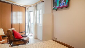 Een hotelkamer met een bed, een stoel met een rood kussen, een aan de muur bevestigde televisie en een raam met gordijnen.
