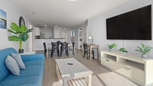 A modern living room with a blue sofa, a coffee table, and a TV on a stand. There is a dining area with a table and chairs, and a kitchen in the background.