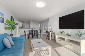 Un salon moderne avec un canapé bleu, une table basse et un téléviseur posé sur un meuble. On aperçoit un coin repas avec une table et des chaises, ainsi qu’une cuisine au fond.