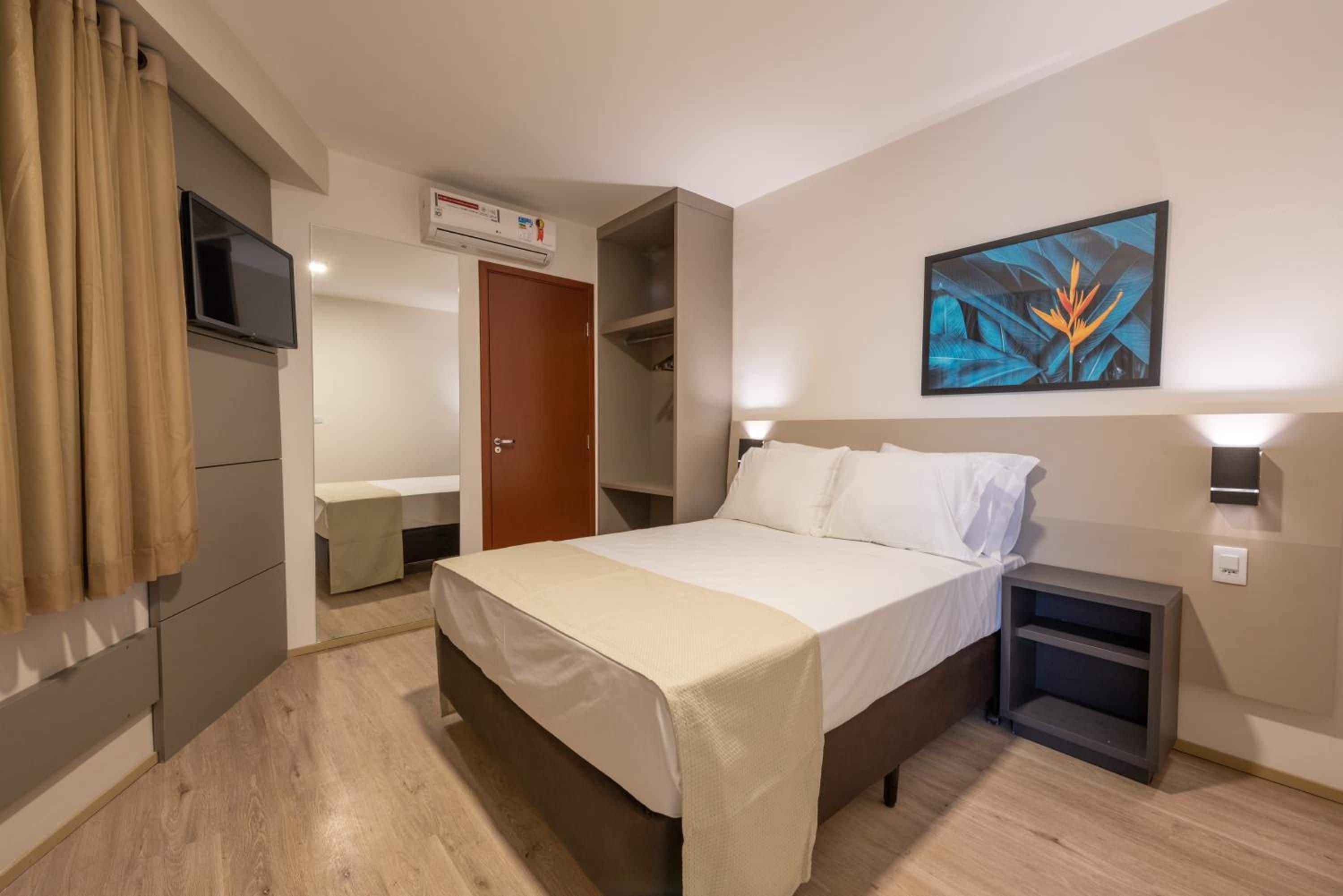 Habitación de hotel con cama, mesitas de noche, televisor, equipo de aire acondicionado y un cuadro en la pared.