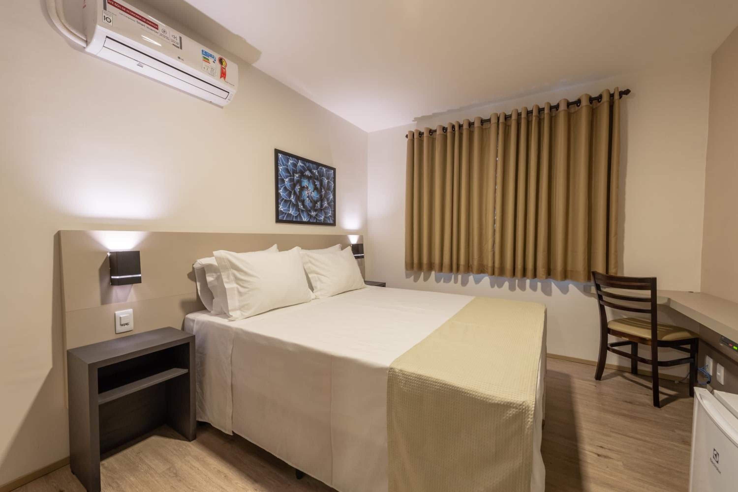 A hotel room with a bed, bedside table, lamp, desk, chair, and an air conditioning unit.