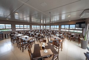 Un comedor amplio con múltiples mesas y sillas, ventanas grandes con vistas a la ciudad y un televisor de pantalla plana en la pared.