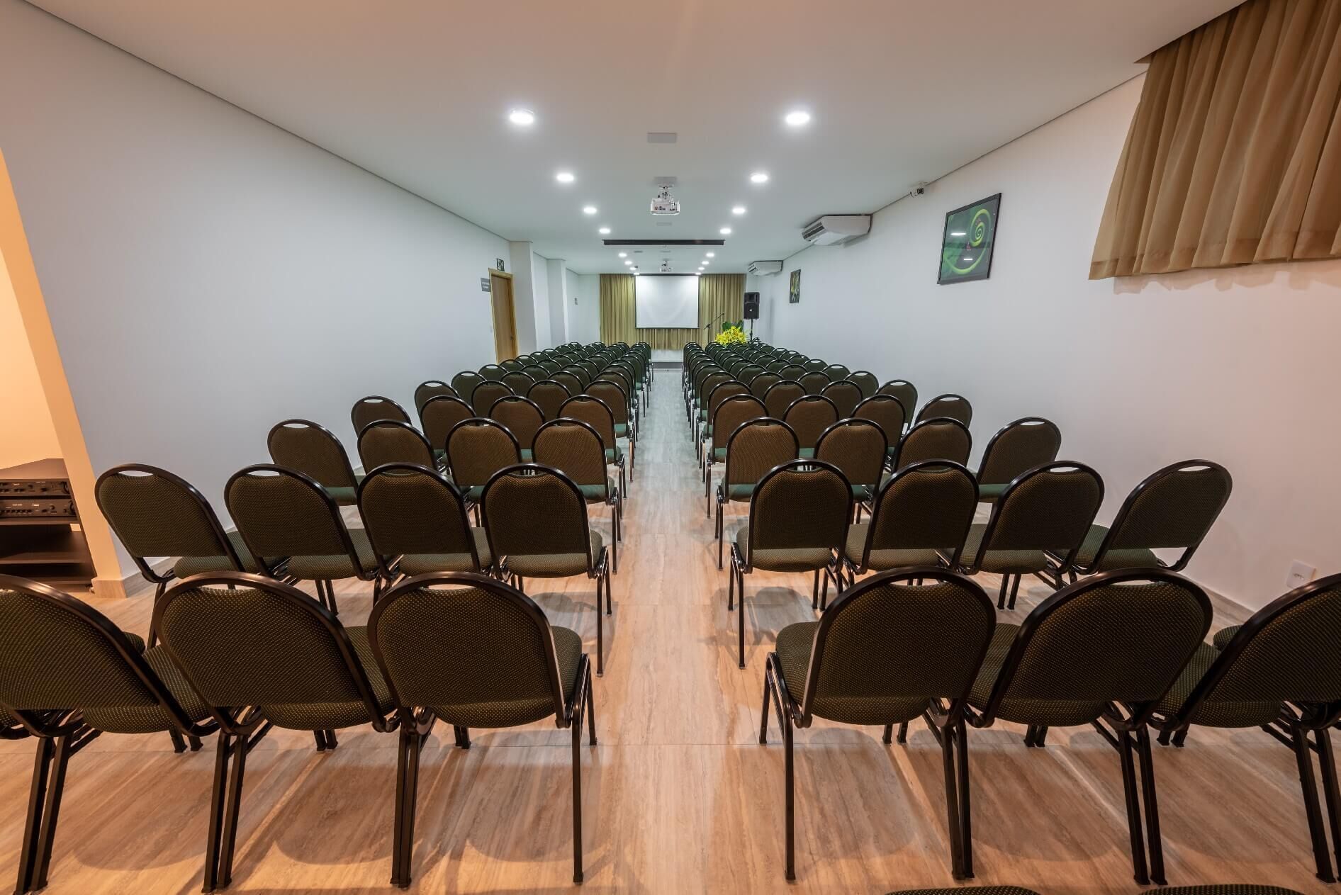 A conference room with rows of chairs facing a stage, equipped with ceiling lights and an air conditioning unit.