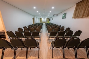 Une salle de conférence avec des rangées de chaises disposées face à une scène, équipée de luminaires au plafond et d’un système de climatisation.