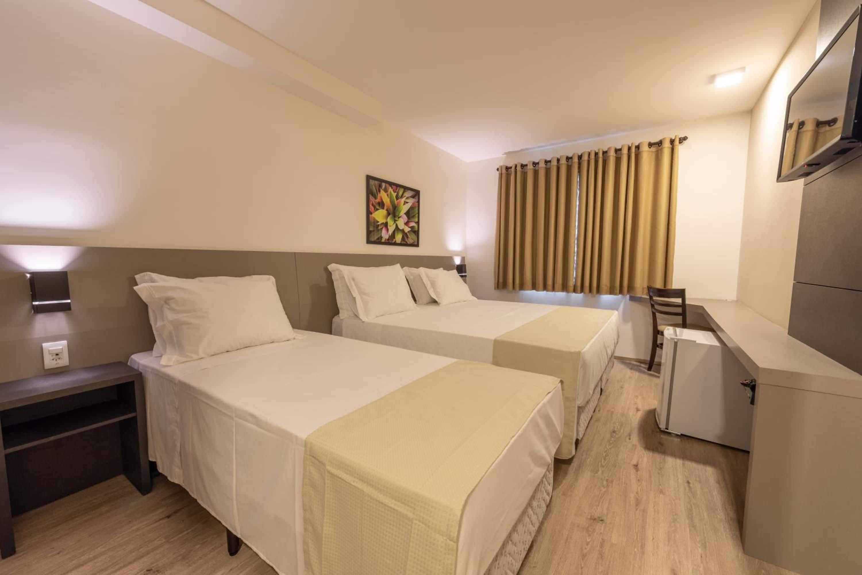 A hotel room with two beds, a desk, a chair, a small refrigerator, and a window with curtains.