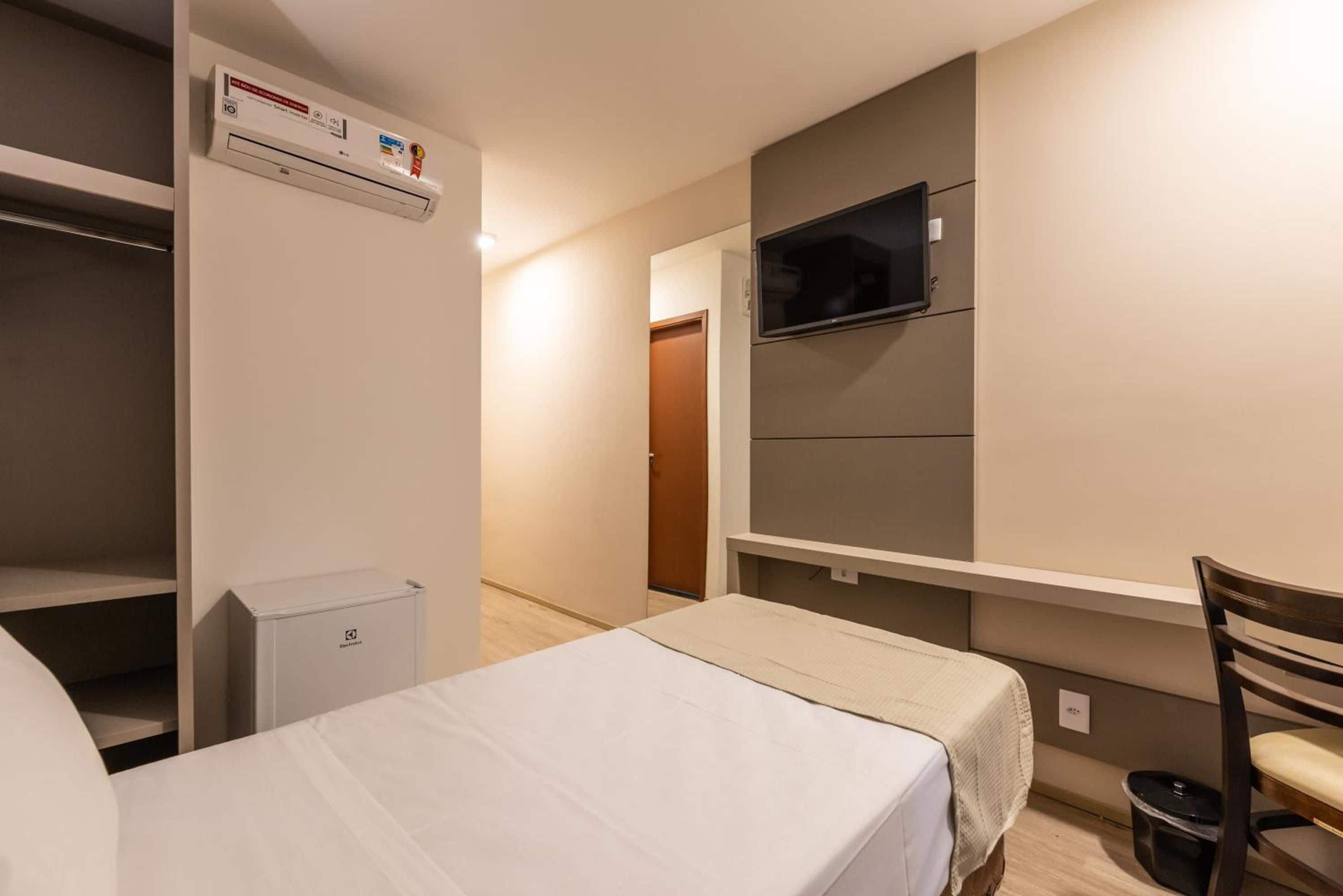 Una habitación de hotel con una cama, un televisor montado en la pared, un refrigerador pequeño y un escritorio con una silla.