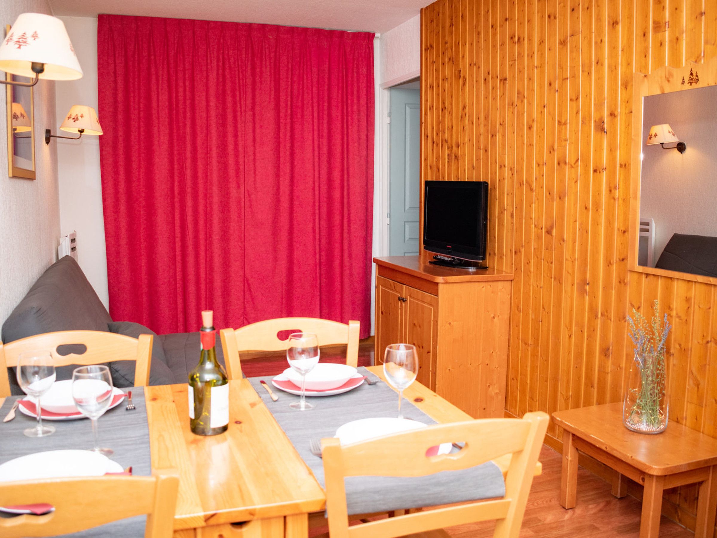A dining area with a wooden table set for two, a television on a wooden cabinet, and a red curtain in the background.