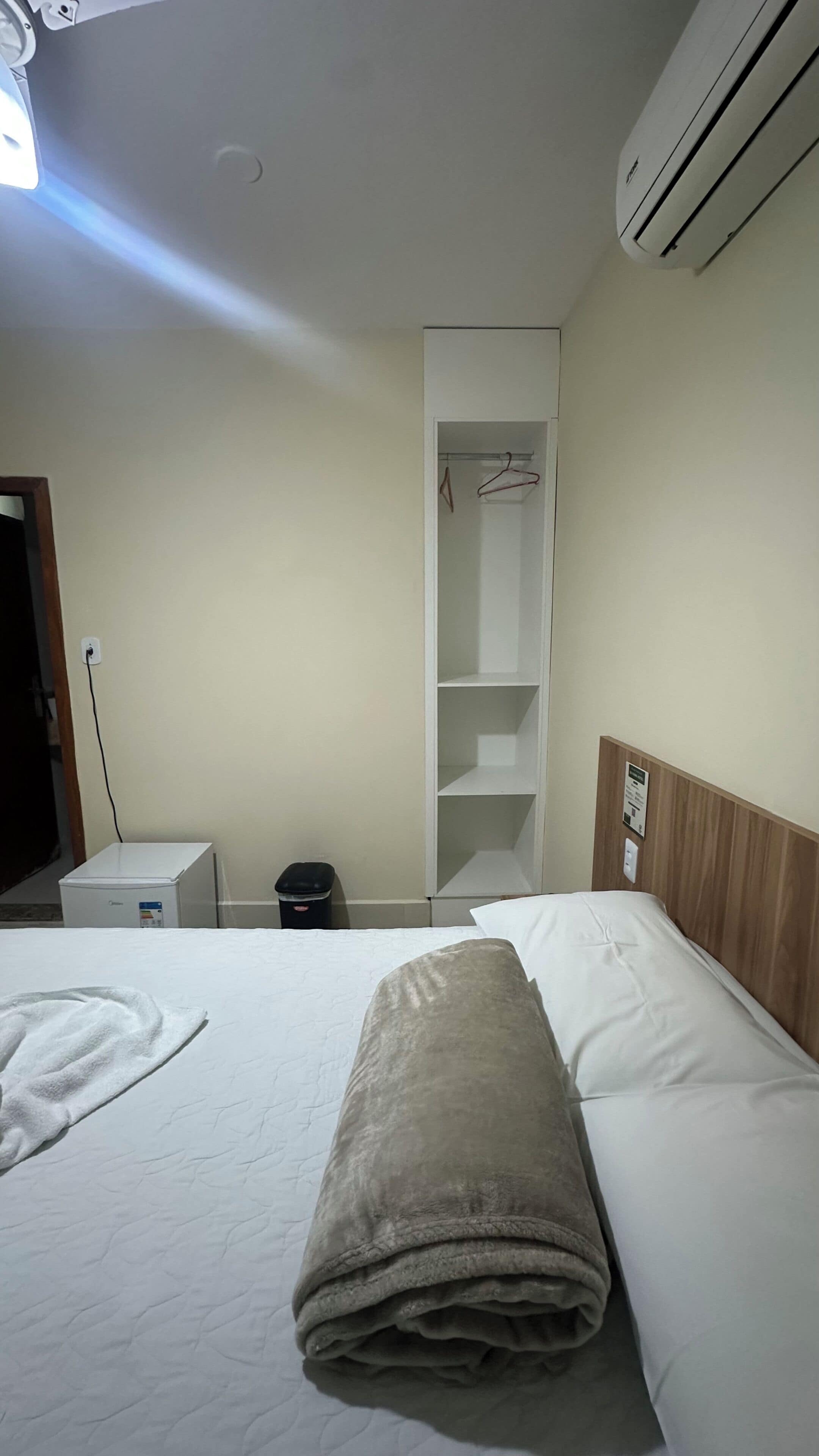 A hotel room with a bed, a white closet, a small table, and an air conditioning unit.