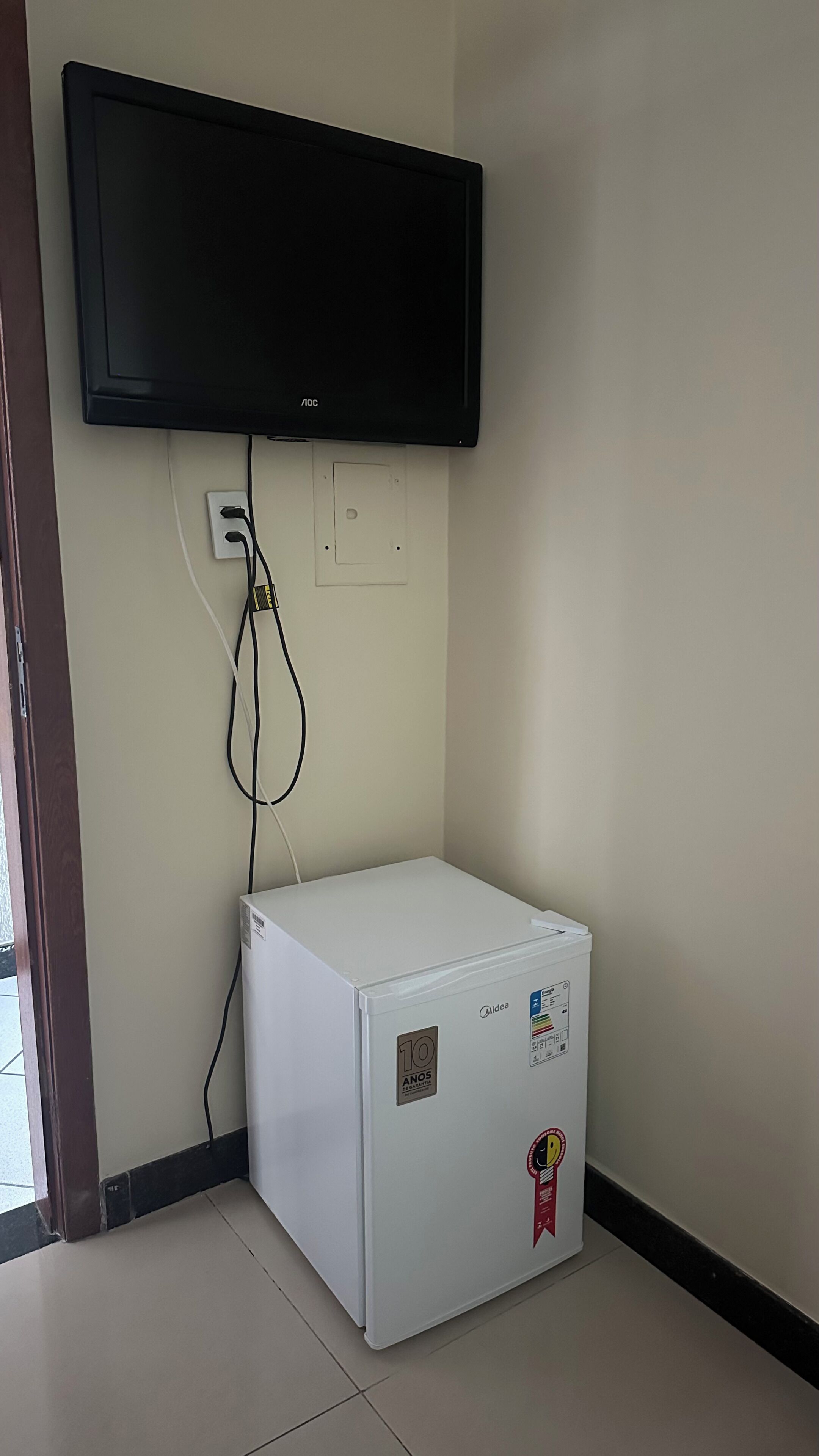 A white refrigerator with a '10 ANOS' sticker, a flat-screen TV mounted above it, and a power outlet on the wall.