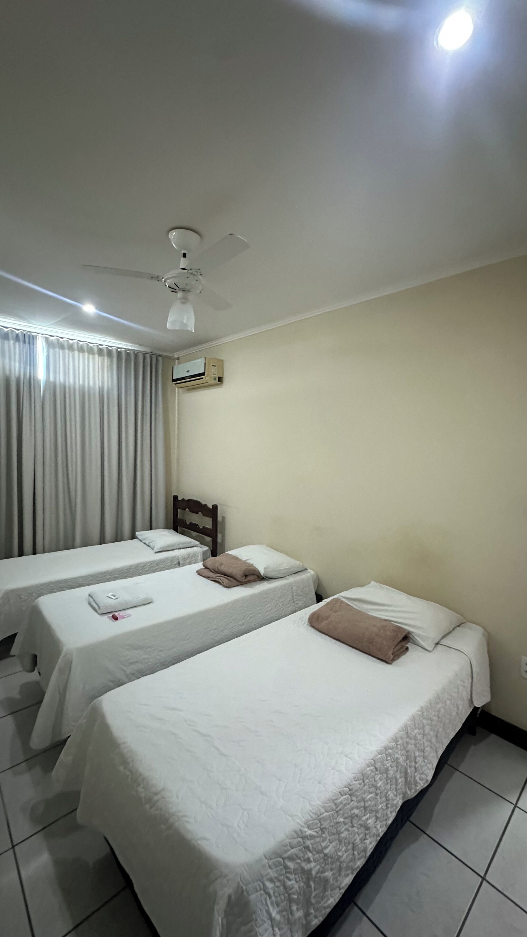 A hotel room with two single beds, a ceiling fan, an air conditioning unit, and a window with curtains.