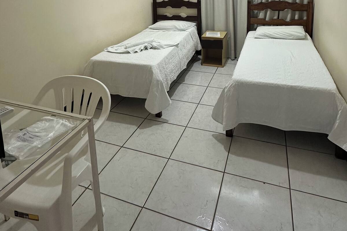 A small, clean room with two single beds, a ceiling fan, an air conditioning unit, a small table with a chair, and a window with curtains.