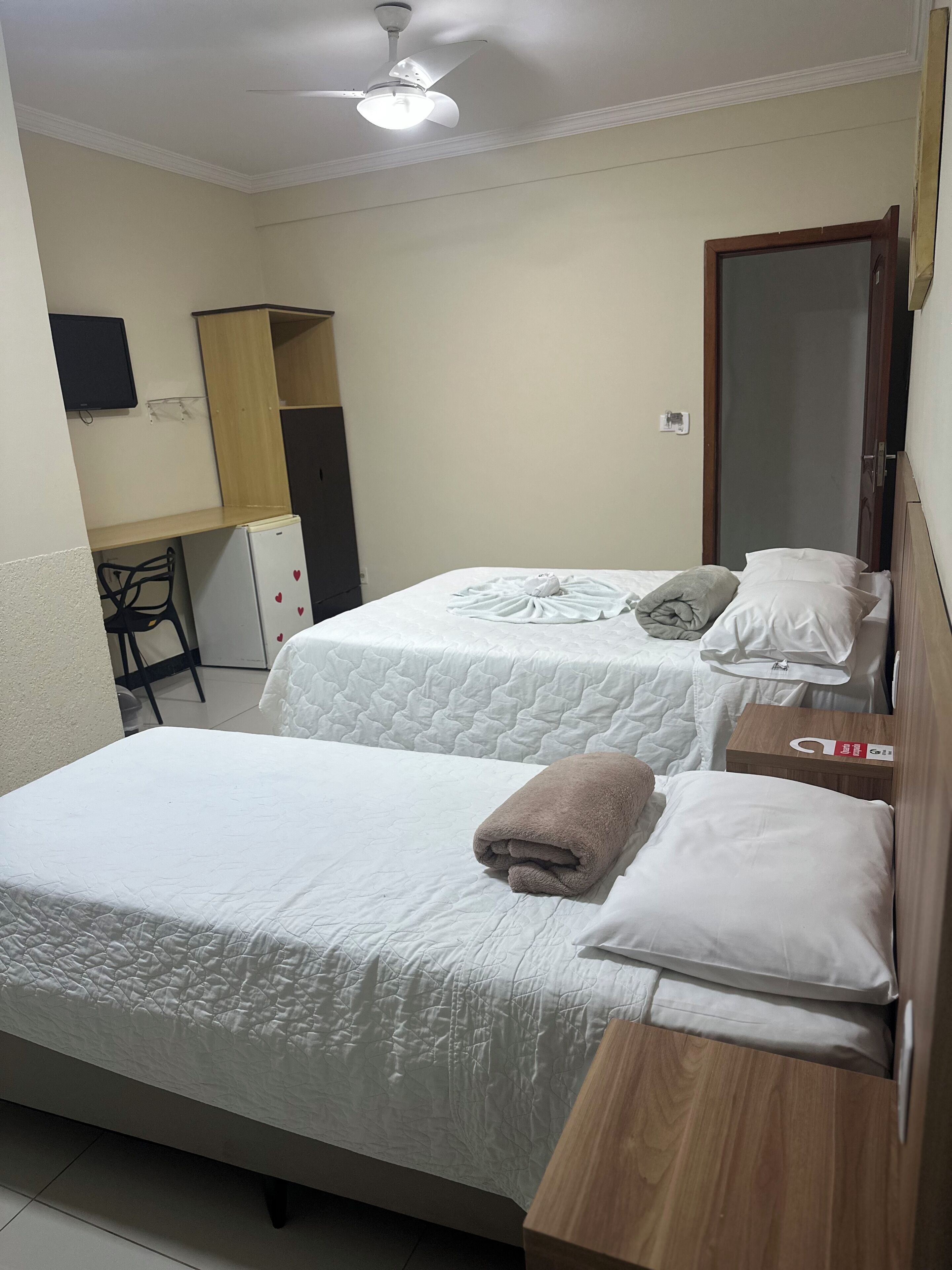 Quarto de hotel com duas camas, uma escrivaninha, uma cadeira, ventoinha no teto e um espelho.